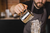 Crop bearded male barista in apron pouring ingredient from stainless pitcher into glass with ice cubes in coffee house