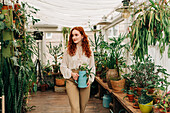 Charming female with long ginger hair standing with jug of seedling in indoor garden with various potted houseplants at home