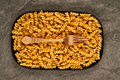 Blick von oben auf eine goldene Gabel, die neben ungekochten Fusilli-Nudeln auf einem Tablett auf dem Tisch liegt