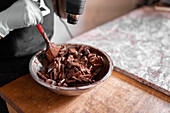 Cropped unrecognizable worker in black apron wearing gloves while melting dark chocolate in stainless steel bowl using heat gun working at counter with marble slab