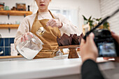 Crop anonymous photographer with photo camera against blogger with jar of icing sugar and muffins in baking cups at home