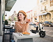 French female in beret sitting at table in cafe with aromatic glass of coffee and freshly baked croissant