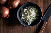 Draufsicht auf eine rustikale Schale mit geschnittenen Zwiebelstücken, die neben einem Messer auf einem Holztisch in der Küche stehen