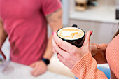 Soft focus of crop unrecognizable female with cup of tasty hot coffee with latte art standing in light kitchen with faceless man
