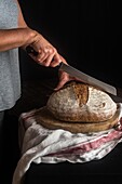 Seitenansicht eines gesichtslosen Kochs mit Brotmesser beim Schneiden eines ganzen Roggensauerteigbrotes auf einem Holzbrett vor schwarzem Hintergrund