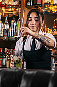 Young brunette in uniform mixing cold cocktail in glass cup while standing behind counter and working in bar