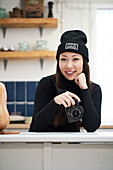 Content ethnic woman with digital photo device leaning with hand on table while looking at camera