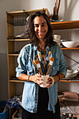 Cheerful adult female master in casual clothes smiling and looking at camera while standing near shelf in workshop with mug of various paintbrushes in hand