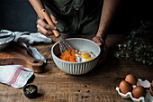 Unrecognizable male cook using whisk to mix pumpkin puree and cream with eggs and flour in bowl while preparing pie on timber table near cutting board and towel