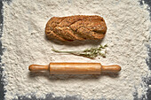 Top view of buckwheat and oat bread with wooden rolling pin and leaves placed on white flour in light room