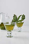 Kleine Gläser mit medizinischem Marihuana-Tee und grünen Kräutern auf weißer Fläche in der Nähe eines Glasgefäßes