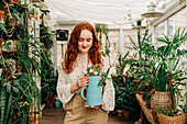 Charming female with long ginger hair standing with jug of seedling in indoor garden with various potted houseplants at home