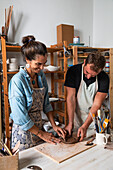 Positive middle aged female and young male in casual clothes and aprons smiling while creating clay plate during pottery class in workshop