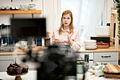 Young female blogger talking against smartphone while recording culinary video and looking at camera at home