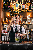 Young female barkeeper pouring alcohol drink from bottle into jigger while preparing cocktail at counter in bar