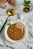 From above of spoon placed near bowl of yummy lentil soup served with slice of sausage and leaf of parsley on marble table and gray napkin