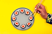 Top view of crop unrecognizable female using bamboo chopsticks while eating delicious sushi rolls with rice and salmon wrapped in nori and served on plate on yellow background