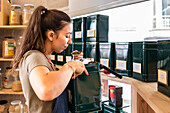 Side view of female seller in apron with ponytail standing near shelves with containers while checking quality of food during working day