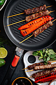 Top view of tasty roasted kebab on skewers and red pepper on grill pan placed on table with assorted vegetables in kitchen
