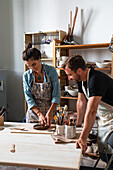 Positive middle aged female and young male in casual clothes and aprons smiling while creating clay plate during pottery class in workshop