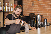 Serious bearded male barista in apron pouring ingredient from stainless pitcher into glass with ice cubes in coffee house