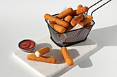 Gebratene Mozzarella-Käse-Sticks in einer Metallfritteuse in der Nähe von Ketchup-Sauce auf einer Marmorplatte auf weißem Hintergrund