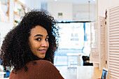 Glückliche Afroamerikanerin mit lockigem Haar sitzt mit Handy am Tresen in einer Bar und schaut über die Schulter in die Kamera