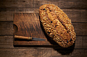 From above of appetizing crispy freshly baked homemade bread loaf with oatmeal flakes and raisin placed with knife on wooden cutting board