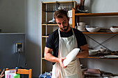 Concentrated young bearded male potter in casual clothes and apron creating white ceramic plate while working in studio