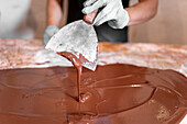 Cropped anonymous employee in gloves using metal spatula to spread liquid chocolate on table while making dessert in confectionery