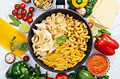 From above of frying pan with various uncooked pasta surrounded with assorted raw vegetables on marble table in light kitchen