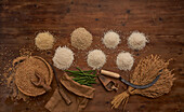 Draufsicht auf kleine Haufen von Bio-Körnern mit frischen Bohnen auf einem Holztisch neben einer Sichel und einem Weizenbündel