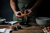 Unrecognizable guy in apron breaking raw egg over bowl while preparing pastry on lumber table near kitchenware