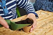 Crop anonymous kid with gardening shovel filling eco cup with earth at table