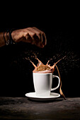 Crop anonymous person throwing tasty sweet cookies into white ceramic cup with splashing coffee on table in kitchen on black background