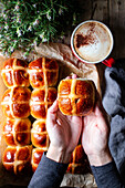 From above crop person hand holding bun on background of table with tasty lush freshly baked hot cross buns and cup of coffee