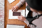 Top view of anonymous woman in sweater and apron placing handmade chocolate bar on piece of paper while working in confectionery