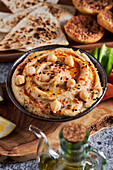 Schale mit leckerem Hummus auf Holzplatte mit verschiedenen Gemüsesorten neben Zitronenscheiben und Ölkrug vor Tortilla-Chips und Croutons