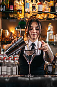 Young tattooed brunette pouring alcohol from shaker through sieve while making tequila sunrise cocktail during work in bar