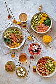 Top view appetizing assorted poke bowls with avocado toasts and glasses of juices served on table with berry dessert near scattered fresh flowers
