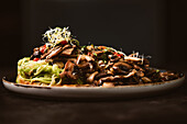 Niedriger Winkel eines leckeren veganen Gerichts mit Zucchini-Spaghetti und sautierten Pilzscheiben, bedeckt mit roten Beeren und Alfalfa-Sprossen auf dunklem Hintergrund
