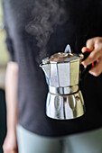 Soft focus of crop unrecognizable person with metal moka pot of steaming hot coffee in hand standing in kitchen at home