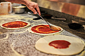 Anonymous man using spoon to smear tomato sauce on pizza bases while working in kitchen of pizzeria