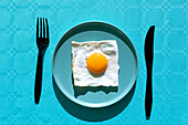 Top view of cooked fried egg place on plate and black fork and knife on light blue background