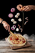 Crop hands breaking egg and spreading sauce and cheese with spatula pizza while mushrooms and onion with bacon falling on top against black background