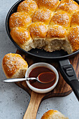 From above of tasty pull apart bread rolls with sesame seeds in frying pan served on wooden cutting board with sweet sauce