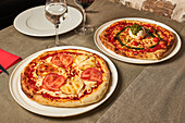 Aufgeschnittene Pizza mit natürlichen Tomatenscheiben und Ziegenkäse neben Pizza mit Kirschtomaten und Parmesan, garniert mit Mozzarella-Käsekugel und Pestosauce, serviert auf einem Teller auf einem Holztisch in einer Pizzeria
