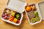 From above of lunch boxes with healthy food including crackers carrot sticks grapes cherry tomatoes with kiwi broccoli walnut and tangerine on yellow background