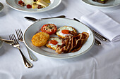 High angle of appetizing fried eggs with bacon slices served on plate with stuffed tomato and crispy mozzarella and placed on table with cutler and cup of coffee during breakfast