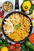 From above of frying pan with various uncooked pasta surrounded with assorted raw vegetables on marble table in light kitchen